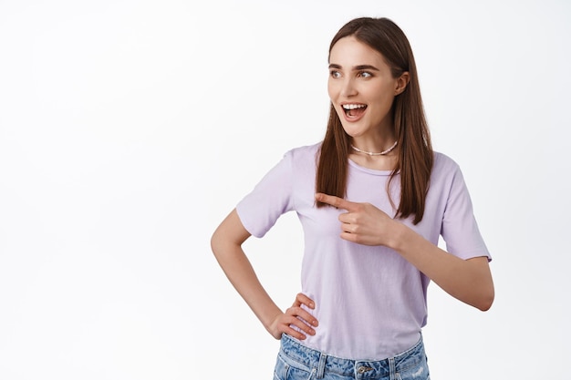 Hey look. Attractive young woman pointing and looking left with excited smile, checking out promo, showing interesting deal in store, intrigued by advertisement text, white background.