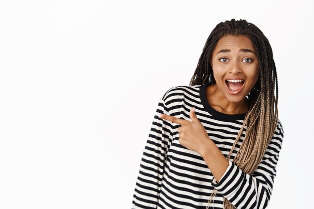 Hey look there Excited Black teenage girl pointing finger away and showing banner smiling happy demonstrating logo or sale offer white background