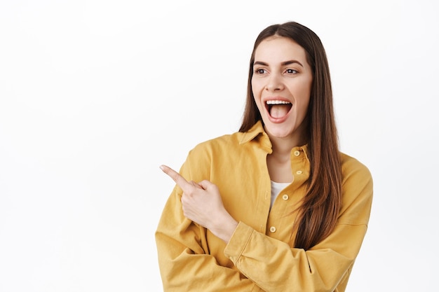 Hey look at this. smiling amused girl checking out awesome deal promo, pointing and looking aside at left copy space, pleased to see something cool, standing over white background