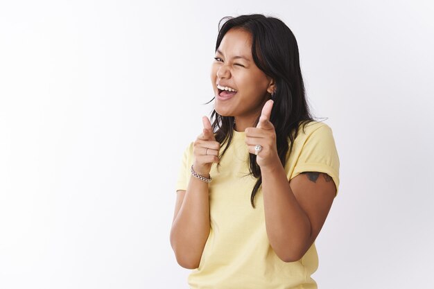 Hey what up. Friendly and outgoing carefree playful polynesian tattooed girl in yellow t-shirt making finger pistols and pointing at camera winking, hinting picking you over white background