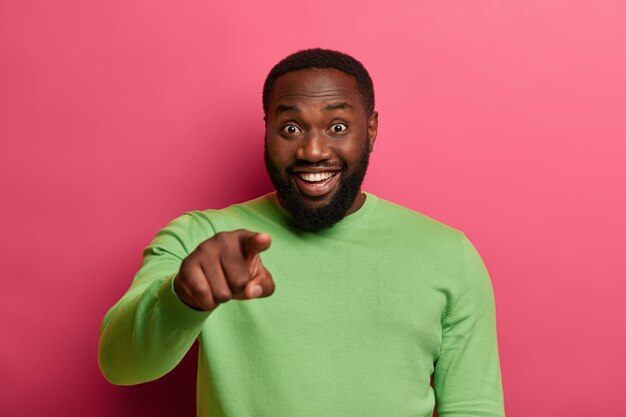 Hey, you. Positive bearded black man points index finger at camera, smiles happily and chooses someone, wears pastel green jumper