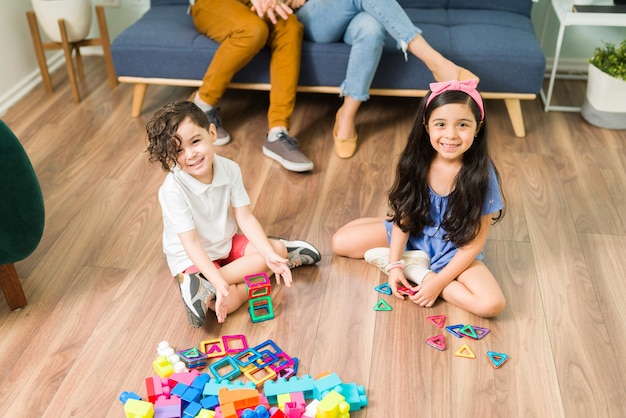 High angle of adorable little siblings smiling while making eye contact and playing a game with fun building blocks in the living room