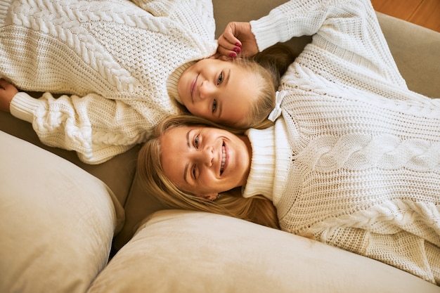 High angle aerial view of overjoyed happy young European mother and her cute little daughter in white jumpers lying comfortably on sofa head to head,  with cheerful smiles