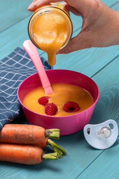 High angle of baby food poured into bowl with carrots and pacifier