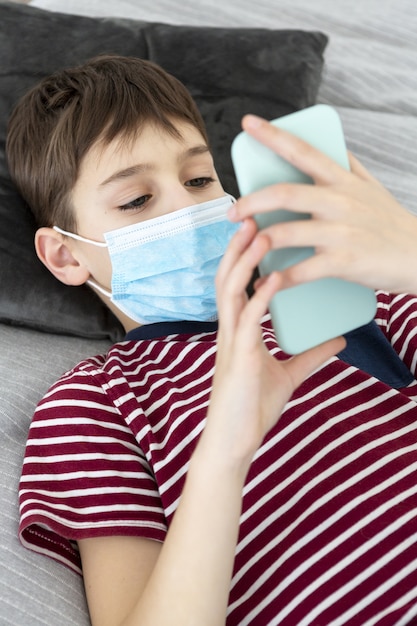 High angle of child with medical mask holding smartphone