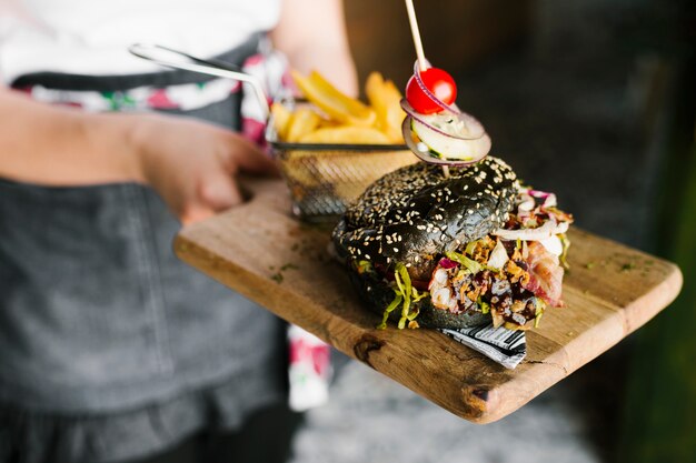 High angle close-up of waiter holding wooden board with black burger and fries