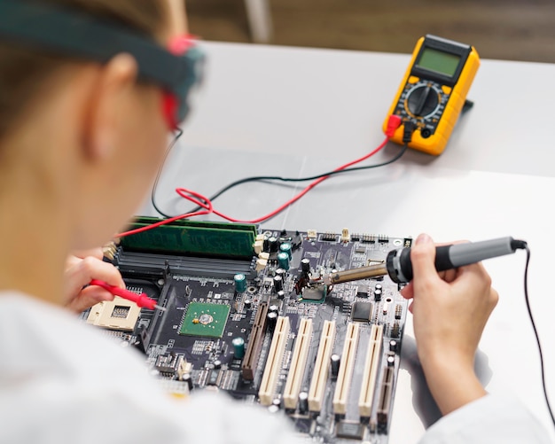 High angle of female technician with soldering iron and electronics motherboard