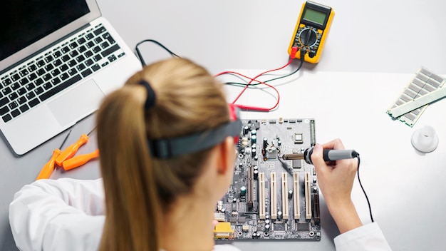 High angle of female technician with soldering iron and electronics motherboard