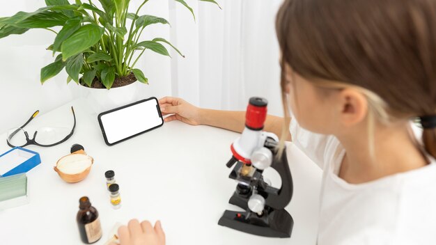 High angle of girl looking into microscope and holding smartphone