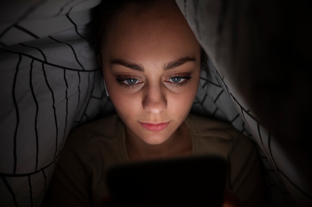 Бесплатное фото Девушка под высоким углом со смартфоном под одеялом