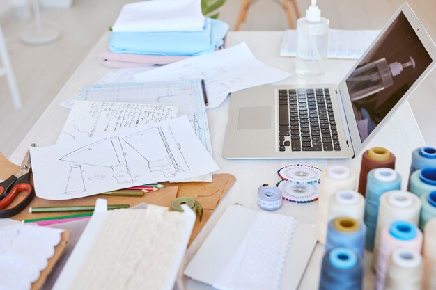 High angle of laptop with clothing line plan and thread spools on table in atelier