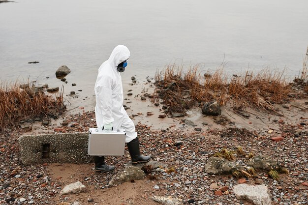 化学​防護服​を​着た​男性​労働者​の​ハイ​アングル​の​肖像画​は​、​屋外​の​産業​廃棄物​の​共同​で​水辺​を​歩いています​。