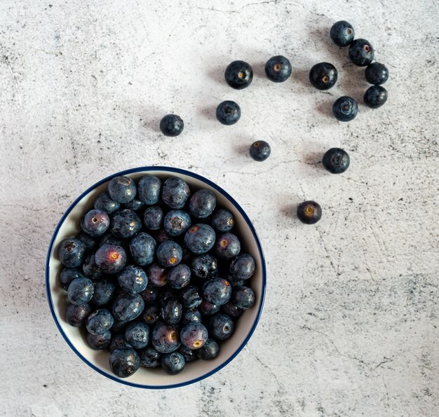 High angle shot of a bowl full of blueberries with some blueberries scattered on a white surface