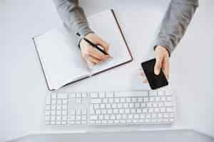 Free photo high angle shot of busy hardworking woman holding smartphone. cropped shot of woman hands making notes in planner and sitting near keyboard and computer, being at work and working with teammates