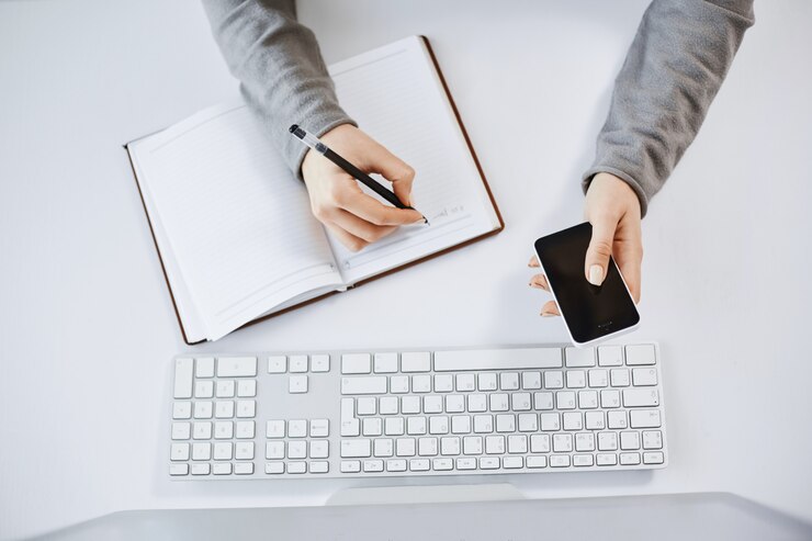 Free Photo high angle shot of busy hardworking woman holding smartphone. cropped shot of woman hands making notes in planner and sitting near keyboard and computer, being at work and working with teammates