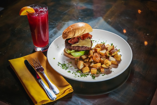 High angle shot of a cheeseburger with potato cubes on the side with a red drink next to it