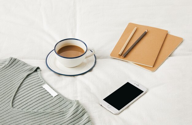 Free Photo high angle shot of a cup of coffee on bedsheets with sketchbooks, phone, and a sweater on it