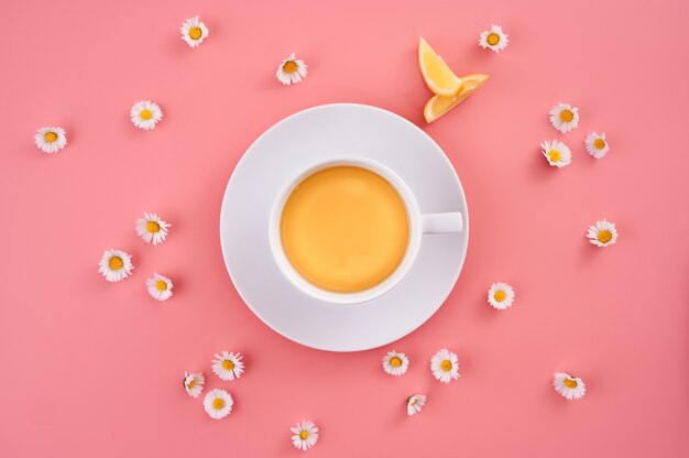 High angle shot of a cup of orange juice surrounded by small daisy flowers on a pink surface