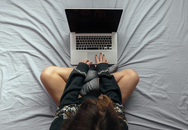 High angle shot of a female typing on the laptop while sitting on the bed with crossed legs