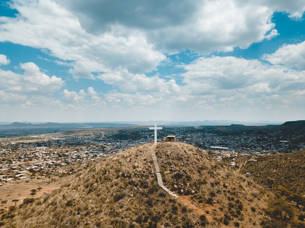 High angle shot of hills covered in dry grass with a big white cross on the top under a blue sky