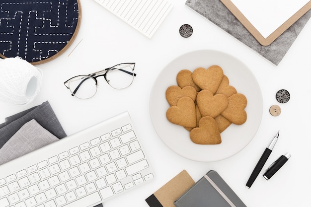 High angle shot of a keyboard, a plate of cookies, glasses, and some papers on a white surface