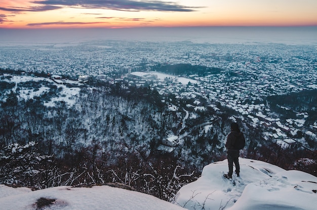 雪に覆われた山の上に立って、街と夕日を眺める男性のハイアングルショット