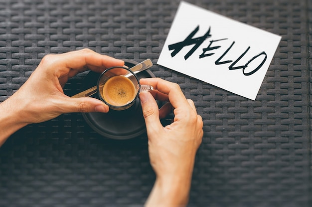 High angle shot of a person drinking a cup of coffee near a hello print on a white card