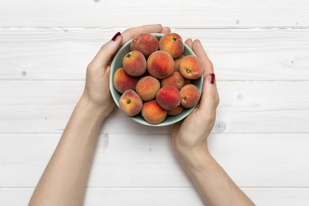 High angle shot of a person holding a green bowl with peaches on a white wooden surface