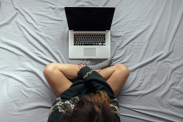 Снимок с высоким углом женщины, работающей с ноутбуком, сидя на кровати со скрещенными ногами