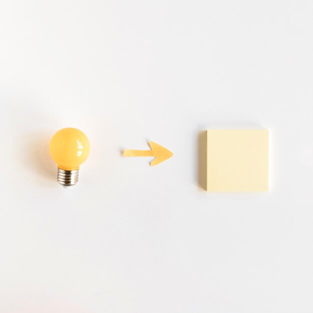 High angle view of arrow symbol between light bulb and adhesive note