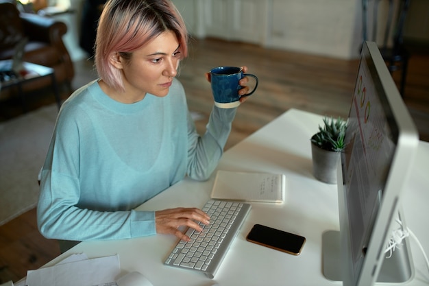 매력적인 젊은 여성 프리랜서의 높은 각도보기는 집에서 멀리 떨어진 곳에서 일하면서 데스크톱 컴퓨터 앞에 앉아 타이핑하고 커피를 마시는 동안 표정을 집중했습니다.