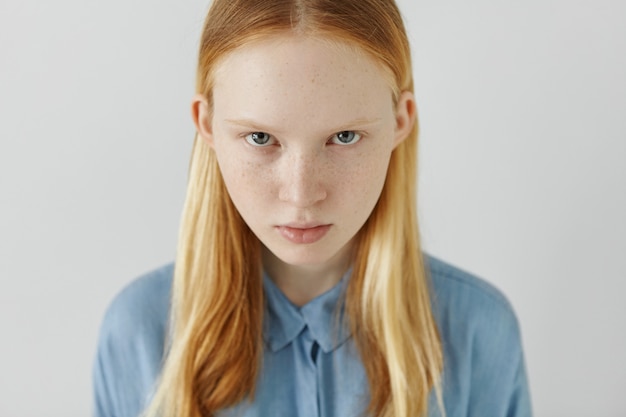 High angle view of beautiful charming woman of tender age staring from under light eyebrows. Pretty girl with freckles wearing her long blonde hair tucked behind ears dressed in blue shirt