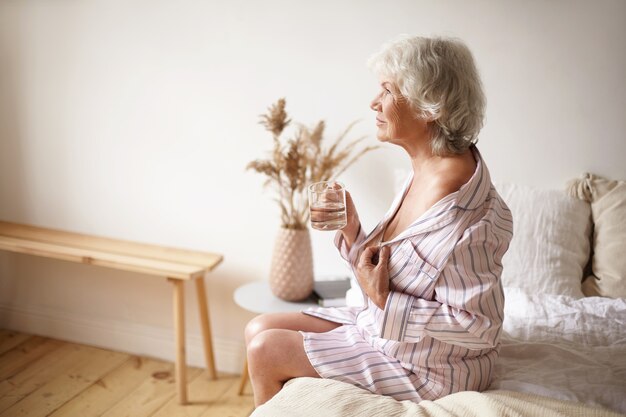 High angle view of beautiful sensual Caucasian sixty year old mature woman in silk pajamas, baring her shoulder while sitting on edge of bed, drinking water after awakening, having happy look