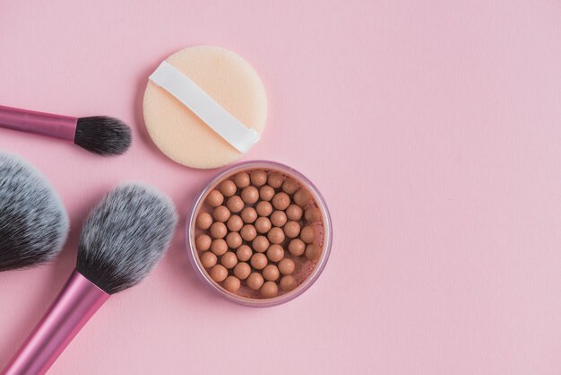 High angle view of bronzing pearls; sponge and makeup brushes on pink surface