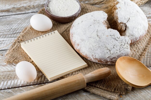 High angle view cake with notepad, eggs, rolling pin on sack cloth and wooden surface. horizontal