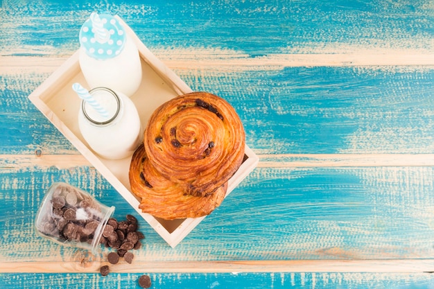 Free photo high angle view of cinnamon bun and milk bottles in wooden tray near spilled choco chips from glass jar