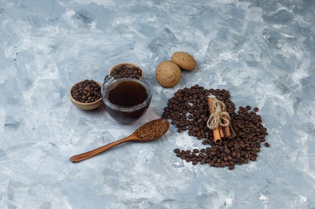 High angle view coffee beans in bowls with cup of coffee, cookies, cinnamon, instant coffee in wooden spoon on light blue marble background. horizontal