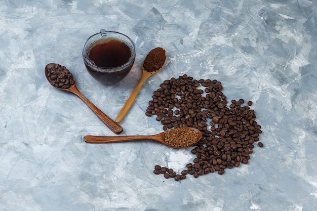 Free photo high angle view coffee beans, cup of coffee with instant coffee, coffee flour, coffee beans in wooden spoons on light blue marble background. horizontal