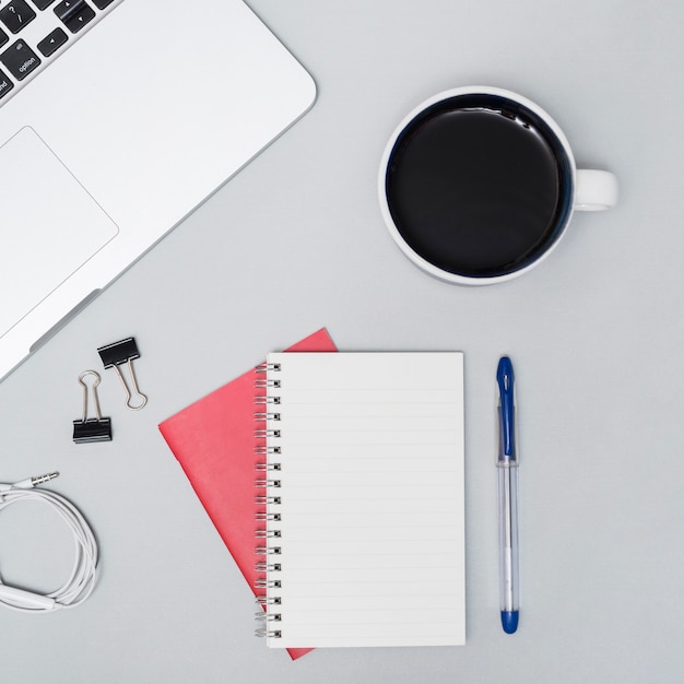 High angle view of coffee cup; laptop; spiral notebook; earphone; pen; against gray background