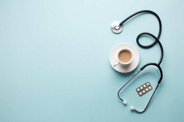 High angle view of coffee cup; stethoscope and medicine in blister pack over blue backdrop