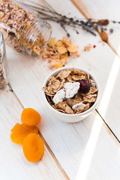 High angle view of cornflakes in bowl near spilled granola and dry fruits on wooden plank