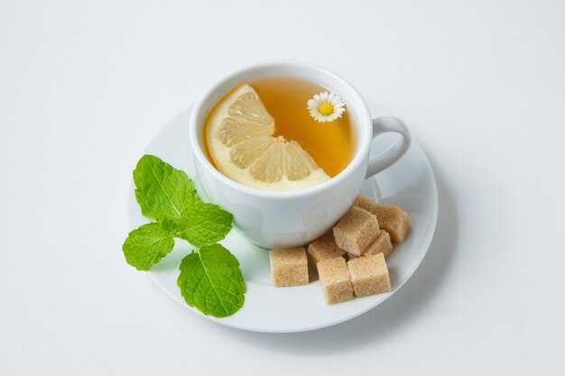 Взгляд высокого угла чашка чая стоцвета с лимоном, листьями мяты, сахаром на белой поверхности. горизонтальный
