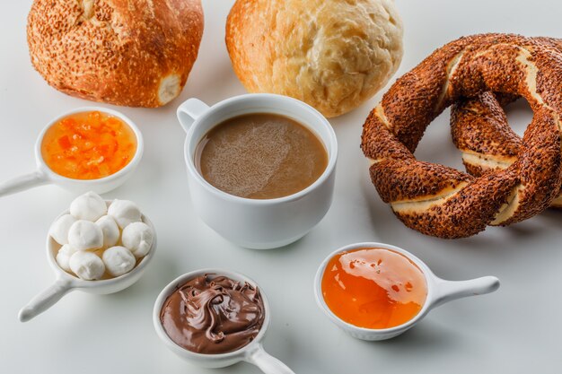 High angle view a cup of coffee with jams, sugar, chocolate in cups, turkish bagel, bread on white surface