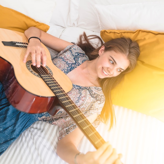 High angle view of happy teenage girl lying on bed playing guitar