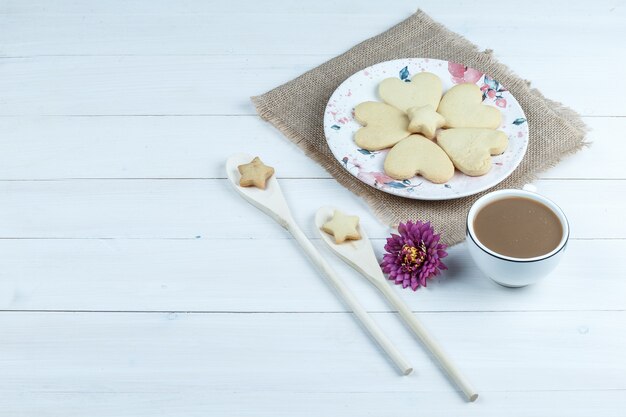 High angle view heart shaped cookies, cup of coffee with flower, star cookies in wooden spoons on white wooden board background. horizontal