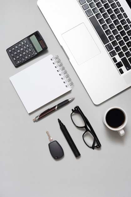 Free photo high angle view of laptop; calculator; blank notepad; eyeglasses; and coffee cup with key on gray backdrop