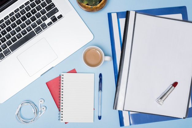 High angle view of laptop; folders; coffee cup; earphone; spiral notepad and pen against blue background