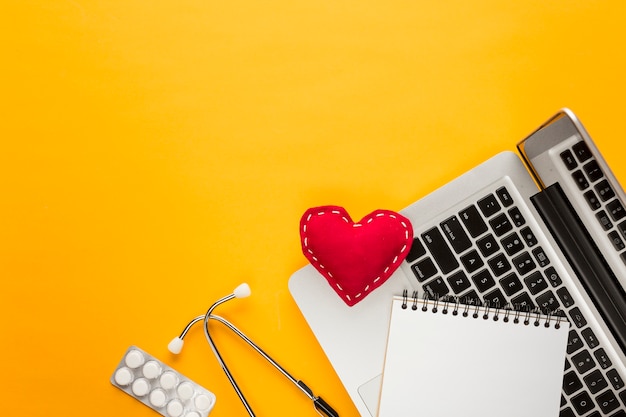 High angle view of laptop; spiral notepad; blister packed medicine; stethoscope; stitched heart shape above yellow backdrop