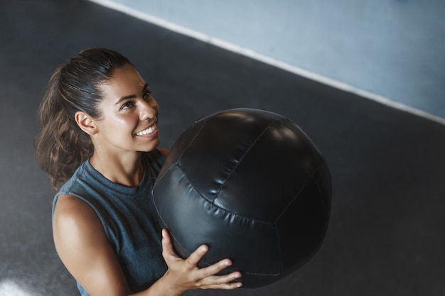 Foto gratuita vista ad alto angolo motivato soddisfatto sudato ispanico sportiva istruttore di fitness sollevare palla medica allenamento crossfit tenere attrezzature per l'allenamento eseguire squat guardando in alto guadagno corpo perfetto