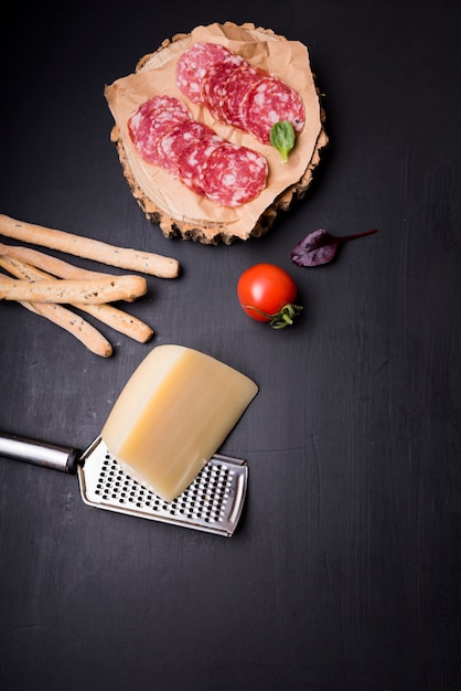 High angle view of pepperoni on wooden coaster with tomato; bread sticks; cheese and grater